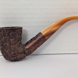 May include: A brown, curved tobacco pipe with a textured bowl and a light brown stem. The pipe has a classic design, with a dark brown, rough-hewn bowl and a smooth, curved stem. The stem is a light amber colour.