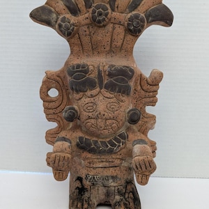 May include: A terracotta-coloured figurine of a stylised figure with a large headdress. The figure has a detailed face, arms, and legs, with darker brown accents. The figurine appears to be handmade and has a textured surface.
