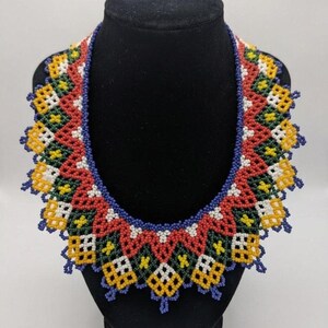 May include: A wide, collar-style necklace crafted from small glass beads in a geometric pattern. The design features repeating zig-zag motifs in red, dark green, yellow, white, and blue, with a scalloped edge and a solid blue beaded border.