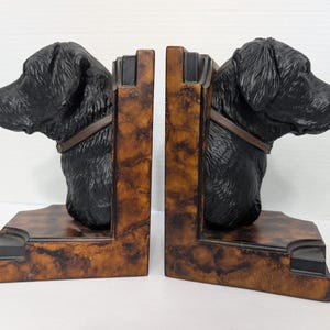 May include: A pair of decorative bookends featuring black dog heads with brown collars. The bookends have a brown and black marbled design and are made of a sturdy material. The dog heads are facing opposite directions.