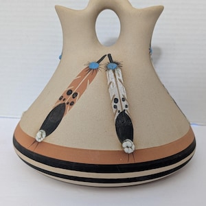 May include: A tan-colored ceramic vase with a unique shape, featuring a handle and two spouts. The vase is adorned with painted feathers in shades of brown and white, accented with black and blue details. A brown and black striped band encircles the base.