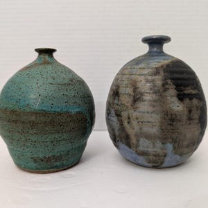 May include: Two ceramic vases. One is teal and brown with a speckled texture. The other is a mix of blue, black, and brown. Both have a rounded body and a narrow neck, suitable for displaying flowers or as decorative pieces.