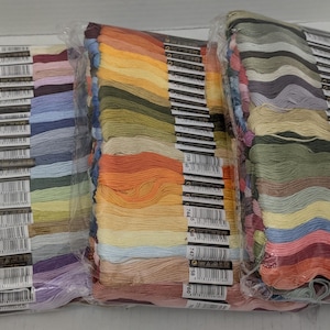 May include: Three clear plastic packages containing bundles of embroidery floss in a wide spectrum of colors. Each bundle features a paper label with a barcode and a unique color number, organized in a vertical row along the side of the thread.