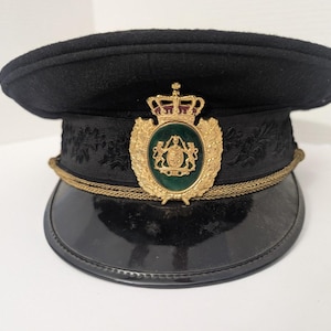 May include: Black military-style hat with a shiny, curved brim. The hat features a gold-colored emblem with a green center and a gold rope trim. The hat has a black, textured crown and a decorative band.