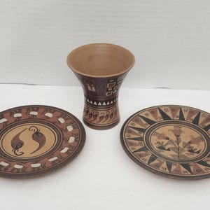 Peut inclure: Un ensemble de trois pièces en céramique comprenant un vase évasé et deux assiettes rondes. Le vase présente des motifs géométriques marron et noirs. Une assiette affiche deux motifs de piments, tandis que l'autre présente un motif floral.