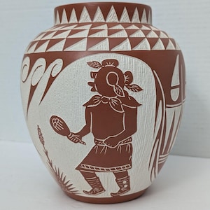 Puede incluir: Un jarrón de cerámica de terracota pintado a mano con un diseño de barbotina blanca. El jarrón presenta una figura estilizada que sostiene una raqueta, rodeada de patrones geométricos y motivos vegetales.