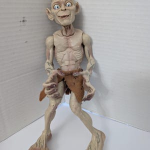 Op de afbeelding: Gedetailleerde actiefiguur van Gollum uit The Lord of the Rings. De figuur is bleek met blauwe ogen, een dun postuur en een versleten lendendoek. De figuur staat licht voorovergebogen met uitgestrekte armen.