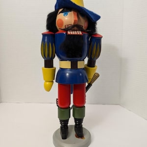 Puede incluir: Figura de cascanueces de madera con uniforme de soldado. La figura tiene un sombrero amarillo, una chaqueta azul, pantalones rojos y botas negras. Tiene barba y bigote negros, y una espada a su lado. El cascanueces se encuentra sobre una base circular gris.