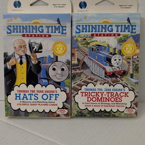 Pode incluir: Duas caixas de jogos de cartas Shining Time Station com Thomas e seus amigos. A caixa da esquerda intitula-se Hats Off, um jogo de memória e correspondência. A caixa da direita intitula-se Tricky-Track Dominoes, um jogo de estratégia.