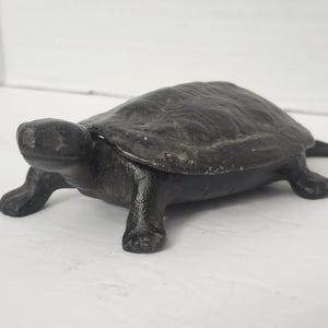 May include: A black cast iron turtle figurine with a textured shell and a hinged lid. The turtle is depicted in a walking position with its head extended forward and four legs planted firmly. The surface of the metal shows a matte finish.