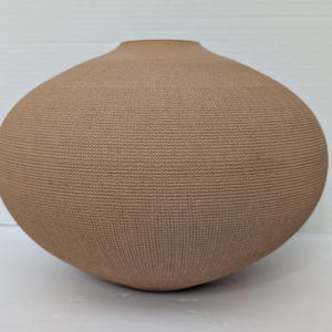 May include: A round, tan-colored ceramic vase with a wide, bulbous body and a small, circular opening at the top. The exterior surface features a fine, uniform grid-like texture that covers the entire rounded form, creating a tactile appearance.