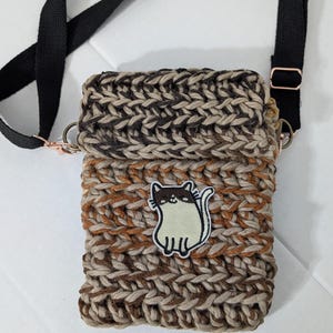 May include: A rectangular crochet crossbody bag featuring a variegated yarn pattern in shades of brown, tan, and cream. The bag has a black adjustable strap with rose gold hardware and a central embroidered patch of a white and brown cat.