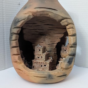 May include: A handcrafted ceramic vase with a unique design. The vase features a cut-out revealing a miniature replica of a stone pueblo village. The exterior is a blend of tan, brown, and grey tones, creating a rustic aesthetic. The vase is approximately 25 cm tall.