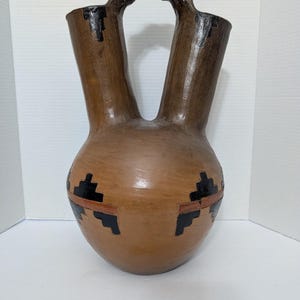 May include: A brown ceramic wedding vase with two spouts connected by a textured handle. The vessel features black geometric patterns around the base and near the top openings. A small rectangular certificate of authenticity is placed in front.