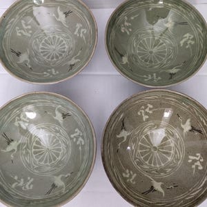 May include: A set of four round ceramic bowls featuring a sage green glaze with white inlaid designs. Each bowl displays a central circular floral motif surrounded by flying crane illustrations and stylized cloud patterns around the interior rim.
