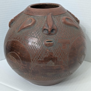 May include: A round, dark brown clay vessel featuring a sculpted human face with a prominent nose and small mouth. The surface is decorated with intricate, etched cross-hatch patterns and geometric shapes that wrap around the entire exterior.