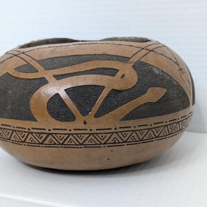 May include: A round, tan-colored gourd bowl featuring a dark brown, textured background with carved, smooth tan snake-like patterns winding around the exterior. A horizontal band with a repeating geometric triangle design encircles the lower section.