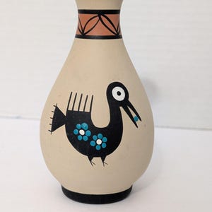 May include: A beige ceramic vase with a black rim and base. The vase features a black bird design with blue flower accents. The neck has a geometric pattern in black and orange. The vase is approximately 6 inches tall.