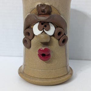 May include: A tan ceramic mug with a face design. The mug features a cowboy hat, large white eyes, a nose, and red lips. The mug has a cylindrical shape with a flared base.