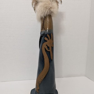 Puede incluir: Un jarrón de cerámica alto con un esmalte azul oscuro y un diseño estilizado marrón. El jarrón tiene una base marrón y una parte superior decorativa con piel y plumas. El jarrón mide aproximadamente 25 cm de alto.