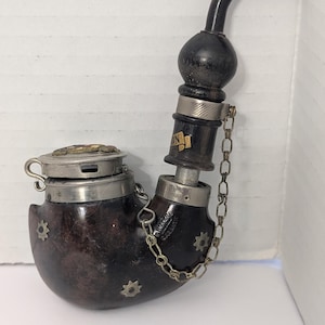 May include: A vintage tobacco pipe with a dark brown bowl and a black stem. The pipe features silver accents, decorative star-shaped inlays, and a hinged lid. A small chain connects the stem to the bowl.