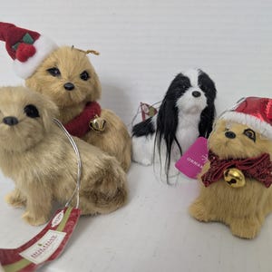 Puede incluir: Tres adornos de perro de peluche que incluyen dos perros de color canela y uno blanco y negro. Un perro canela lleva un gorro de Papá Noel rojo, el otro lleva un gorro con un lazo y un cascabel, y el perro blanco y negro tiene pelo largo.