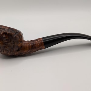 May include: A tobacco pipe featuring a dark brown, textured bowl and a curved, black stem. The bowl has a carved design, and the stem is joined to the bowl with a lighter brown band. The pipe is set against a white backdrop.