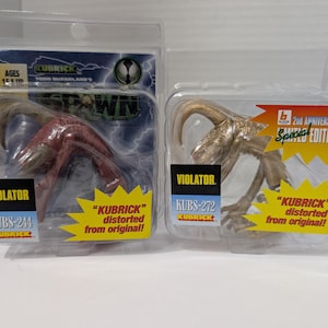 Op de afbeelding: Twee "Kubrick" actiefiguren van het personage "Violator" uit de "Spawn" stripreeks. Eén figuur is rood, de andere is bruin. Beide zitten in een transparante plastic verpakking met het "Spawn" logo en de tekst "2nd Anniversary Special Edition".