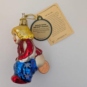 May include: A glass Christmas ornament in the shape of a child, with blonde hair and a red, white, and blue outfit. The ornament has a gold metal cap and a tag that reads "Authentic German INGE-GLAS Ornaments."