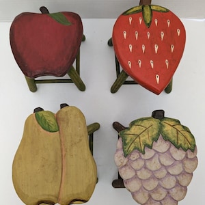 May include: Four wooden stools painted to look like fruit. The stools are painted to look like an apple, a strawberry, a pear, and a bunch of grapes. The stools are all on a white surface.