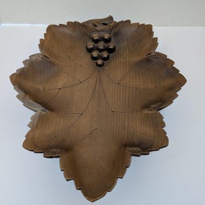 Puede incluir: Un cuenco de madera marrón con forma de hoja y detalles tallados. El cuenco presenta un racimo de uvas en el centro y intrincados patrones de vetas. Los bordes del cuenco son festoneados, lo que aumenta su atractivo decorativo. El cuenco es de madera.