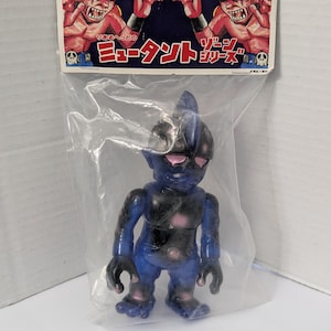 May include: A blue and black plastic toy figure of a monster, packaged in a clear plastic bag. The figure has a fin on its head and pink eyes. The packaging features illustrations of red monsters and Japanese text.