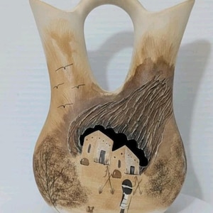 May include: A cream-colored, double-spouted vase with a central handle. The vase features a painted landscape scene with two buildings, a figure, trees, and birds. The design incorporates shades of brown and tan, with a textured effect on the background.