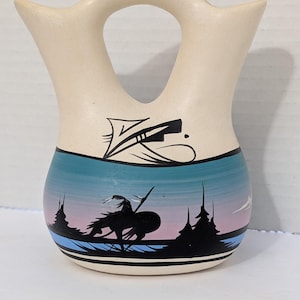 May include: A cream-coloured ceramic vase with a double spout and a handle. The vase features a painted landscape scene with a Native American rider on horseback, silhouetted against a blue and pink sky. Black geometric designs are painted near the top.