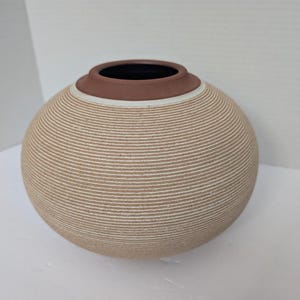 May include: A spherical ceramic vase with a textured surface featuring fine horizontal ridges in alternating shades of beige and white. The vase has a smooth, dark brown circular rim at the top opening, contrasting with the light-colored body.