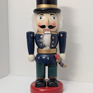 Puede incluir: Figura de cascanueces de madera con uniforme de estilo militar. La figura tiene un sombrero de copa negro con ribete dorado, barba blanca y chaqueta azul con detalles dorados. Se apoya sobre una base roja y sostiene una espada pequeña.