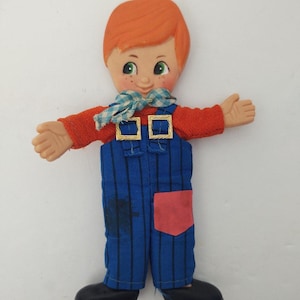 May include: A vintage doll figurine of a boy with orange hair, green eyes, and freckles. He wears a red sweater, blue overalls with gold buckles, and a blue and white checkered scarf. The doll has black shoes and outstretched arms.