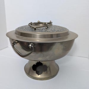 Peut inclure: Un plat à réchaud argenté métallique avec un bol large, deux poignées latérales et une base sur pied dotée d'une découpe à quatre lobes. Le couvercle possède une poignée centrale et une surface supérieure décorée d'un motif circulaire.