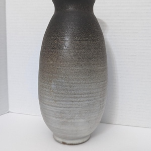 May include: A handcrafted ceramic vase with a gradient color scheme. The top is dark brown, transitioning to gray and then white at the base. The vase has a rounded body and a narrow neck, perfect for displaying flowers or as a decorative piece.