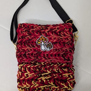 May include: A rectangular crochet crossbody bag featuring a thick, textured knit pattern in shades of red, burgundy, and yellow. The bag includes a black adjustable strap with gold-tone metal hardware and a small mushroom patch on the front.