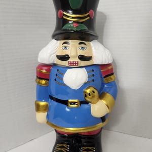 Peut inclure: Une figurine de casse-noisette en céramique avec un chapeau noir orné d'un panache vert, des cheveux blancs et un uniforme bleu aux détails dorés. Le personnage porte une moustache noire, une barbe blanche et des bottes noires à lacets jaunes.
