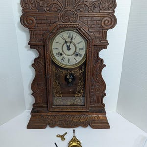 May include: A vintage wooden clock with ornate carvings and a glass front. The clock face is white with black Roman numerals. Includes a key, pendulum, and a small tool. The clock is brown and has a decorative top.
