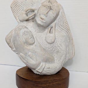 以下が含まれることがあります： 母と子の彫刻が施された石の彫刻。彫刻は明るい灰色で、丸い濃い木製の台座に取り付けられています。母親の姿は子供を抱いており、両方の姿が正面を向いています。