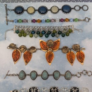 May include: Six distinct bracelets arranged vertically. The first features dark blue and cream circular stones. The second has multi-colored crystal beads. The third is a silver chain with dangling green beads. The fourth has amber leaf charms and metal flowers. The fifth has turquoise teardrop stones. The sixth has black rectangular stones in silver frames.