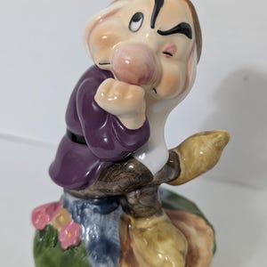 Könnte beinhalten: Eine Keramikfigur des Charakters Grumpy aus Schneewittchen, dargestellt mit gerunzelter Stirn, einer großen rosa Nase und einer violetten Tunika. Die Figur sitzt auf einem Sockel mit grünem Gras, kleinen rosa Blumen und braunen Felsstrukturen.