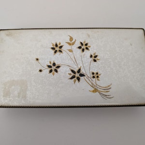 May include: Rectangular white trinket box with a floral design. The box features a speckled surface and a decorative arrangement of black and gold flowers. The edges are trimmed with a dark metal border, creating a vintage aesthetic.