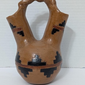 May include: A brown pottery vessel with two spouts connected by a handle. The vessel is decorated with black and red geometric patterns. The pottery has a rounded base and a textured surface.