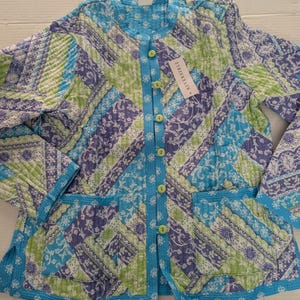 May include: Reversible quilted jacket in a patchwork design. The jacket features a mix of blue, green, and purple floral patterns. It has a round collar, button closure, and two front pockets. The jacket has a tag that says "Reversible".