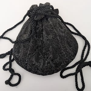 Può includere: Una borsa nera con coulisse e perline con un motivo strutturato. La borsa ha una forma arrotondata ed è adornata da una spessa corda nera ritorta che funge da coulisse e tracolla. La borsa è su uno sfondo bianco.