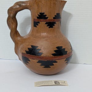 May include: A brown ceramic pitcher with a unique handle and a spout. The pitcher features a design of black geometric shapes and red bands around the body. The handle has a textured, branch-like appearance. A small tag is visible.